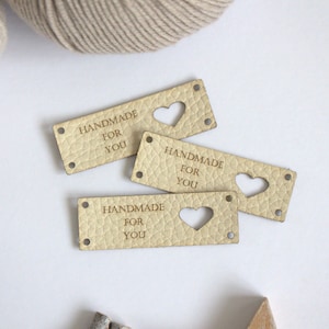 May include: Four rectangular, light tan leather tags with the text "HANDMADE FOR YOU" engraved. Two tags have a heart-shaped cutout. Each tag has four small holes for sewing. Beige yarn is in the background.