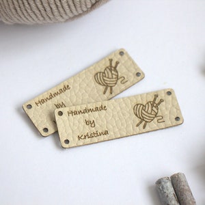 May include: Two beige leather tags with the text "Handmade by Kristina" and a heart-shaped yarn design. The tags have small holes at each end. A ball of yarn and decorative wooden beads are in the background.