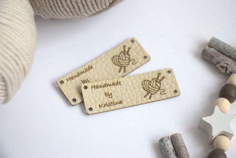 May include: Two beige leather tags with the text "Handmade by Kristina" and a heart-shaped yarn design. The tags have small holes at each end. A ball of yarn and decorative wooden beads are in the background.