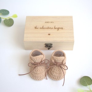 Könnte beinhalten: Ein Paar hellbraune, gehäkelte Babyschuhe mit braunen, lederartigen Schnürsenkeln vor einer rechteckigen Holzschatulle. Auf dem Deckel der Schatulle befindet sich der eingravierte Text "AND SO, the adventure begins..." mit einem kleinen Herzsymbol.