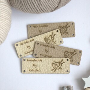 May include: A collection of beige leather tags with the words "Handmade by Kristina" and a graphic of yarn and knitting needles. The tags have two holes for sewing and are arranged on a white surface with yarn and decorative elements.