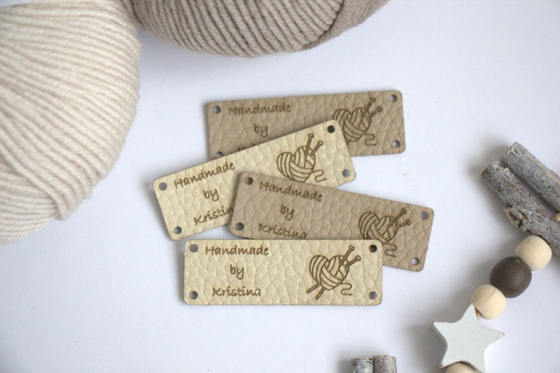 May include: A collection of beige leather tags with the words "Handmade by Kristina" and a graphic of yarn and knitting needles. The tags have two holes for sewing and are arranged on a white surface with yarn and decorative elements.