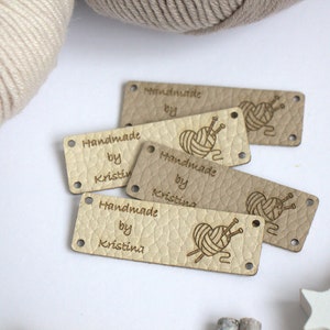 May include: A collection of beige leather tags with the text "Handmade by Kristin" and a heart-shaped yarn and needle design. The tags have small holes for sewing and are arranged on a white surface with yarn and decorative elements.