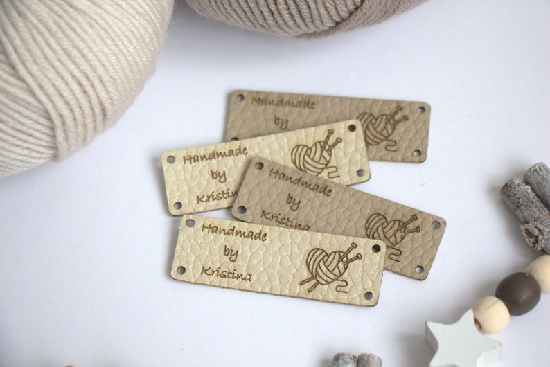 May include: A collection of beige leather tags with the text "Handmade by Kristin" and a heart-shaped yarn and needle design. The tags have small holes for sewing and are arranged on a white surface with yarn and decorative elements.