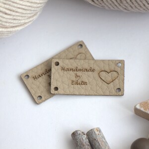 May include: Two beige leather tags with the text "Handmade by Edita" and a heart symbol. The tags have four holes for sewing and are placed on a white surface with yarn and wooden decorative elements.