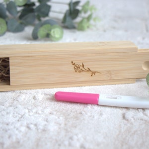 Könnte beinhalten: Eine hellfarbene Bambusbox mit Schiebedeckel, verziert mit einem floralen Design. Ein rosa-weißer Schwangerschaftstest liegt vor der Box. Die Box enthält geschreddertes Papier.