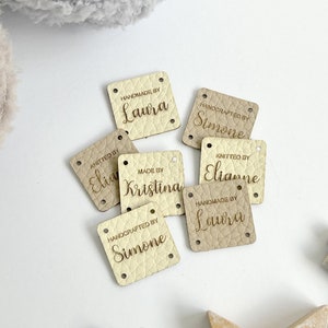 May include: Several square, off-white leather tags with rounded corners. Each tag has a name and the phrases "Handmade by," "Knitted by," or "Handcrafted by" in a light brown font. These tags are for handmade crafts.