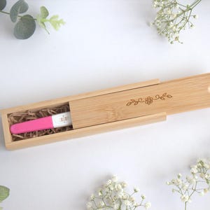 Könnte beinhalten: Ein rosa Schwangerschaftstest liegt in einer Holzbox mit Schiebedeckel, verziert mit einem eingravierten Blumendesign. Der Hintergrund ist weiß, mit Zweigen weißer Blumen und Grün.