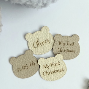 May include: Four bear-shaped tags in beige tones. One tag says "Oliver", another "My First Christmas", and a third "11.05.24". The tags have a textured surface, possibly leather, and are likely decorative or personalized items.