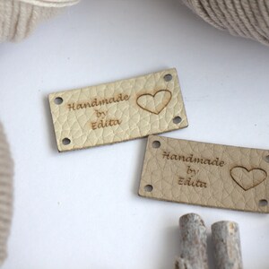 Puede incluir: Dos etiquetas rectangulares de cuero color beige claro con el texto "Handmade by Edita" y un símbolo de corazón. Cada etiqueta tiene cuatro pequeños agujeros en las esquinas. Las etiquetas están sobre una superficie blanca, con hilo y palitos de madera en el fondo.
