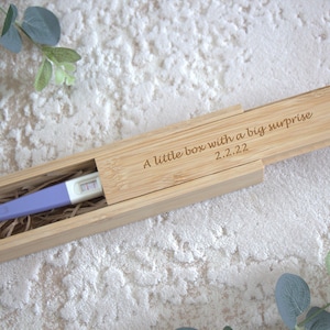 May include: A bamboo box with a sliding lid, holding a positive pregnancy test. The box is engraved with the words "A little box with a big surprise 2.2.22". The test has a purple handle.