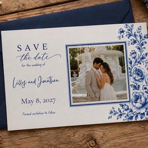 May include: A rectangular Save the Date card featuring blue floral illustrations on the right side and a central photograph of a couple in front of a fountain. The text reads: SAVE the date for the wedding of Lilly and Jonathan May 8, 2027.