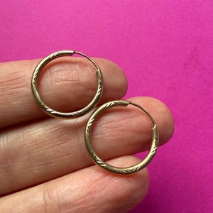 May include: A pair of gold hoop earrings featuring a delicate, repeating etched pattern along the outer surface of the circular bands. The earrings are constructed from a thin, polished gold wire that forms a continuous, lightweight circular shape.