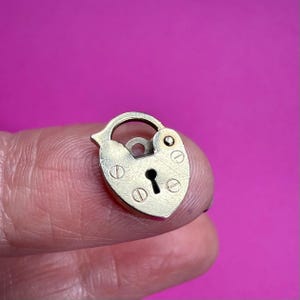 May include: A small, heart-shaped brass padlock with a keyhole and a curved handle. The padlock has a vintage aesthetic, with visible screw heads and a tarnished finish. The background is a solid magenta color.