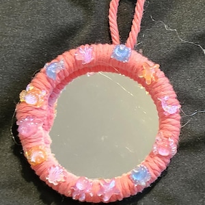 May include: A circular mirror with a frame wrapped in pink yarn, featuring a pink yarn loop at the top for hanging. The frame is decorated with small, translucent, multi-colored plastic charms shaped like sea creatures, stars, and shells.