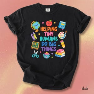 May include: A black short-sleeve t-shirt featuring the text HELPING TINY HUMANS DO BIG THINGS in colorful letters, surrounded by illustrations of a globe, apple, book, crayons, paint palette, chalkboard, pencil, scissors, sippy cup, and books.