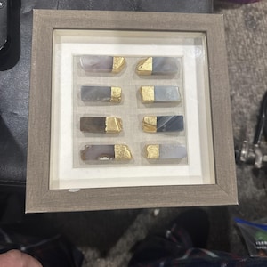 May include: A square, light brown wooden frame containing eight rectangular, polished agate stone pieces arranged in two vertical columns. Each stone features a natural grey or brown banded pattern and is tipped with a textured gold-colored accent.