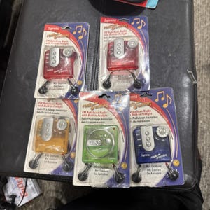 Puede incluir: Cinco radios FM AutoScan Suprema Radio Action con linterna integrada, cada una en su empaque blíster original. Las radios vienen en rojo, naranja, verde y azul, e incluyen auriculares con cable negros. Cada empaque muestra el nombre del producto y texto.