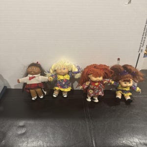 May include: Four dolls with yarn hair and fabric clothing. The first has dark hair, a white top, and a red plaid skirt. The second has blonde hair and a floral dress. The third has red hair and a floral dress. The fourth has auburn hair and a floral dress.