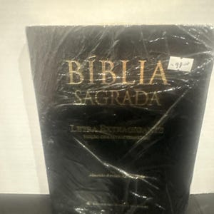 May include: A black hardback book titled BÍBLIA SAGRADA with gold lettering on the front cover. The text includes LETRA EXTRAGIGANTE and EDIÇÃO COM LETRAS VERMELHAS. The book is wrapped in clear plastic film and features a small white price tag.
