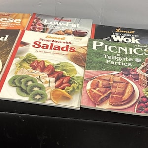May include: A collection of seven Sunset cookbooks featuring titles such as Seafood, Cheese, Low-Fat, Fresh Ways with Salads, The Complete Wok, Picnics & Tailgate Parties, and Barbecue, each displaying colourful food photography on the front covers.