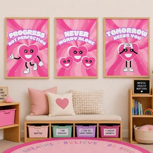 Pode incluir: Três quadros de parede com personagens de corações cor-de-rosa com rostos sorridentes e fundos ondulados. Os quadros exibem os textos Progress Not Perfection, Never Worry Alone e Tomorrow Needs You em letras brancas arredondadas.