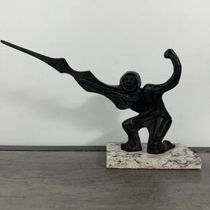 May include: A black sculpture of a figure in a kneeling position, with an arm raised and a long, wing-like element extending from the back. The sculpture is mounted on a rectangular base with a speckled pattern.