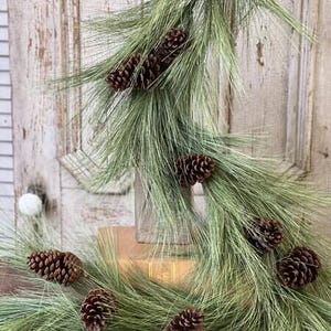 May include: A decorative garland featuring long, slender green pine needles and multiple brown pinecones. The garland is draped over a rectangular wooden block, showcasing a natural, textured appearance with varied pinecone sizes.