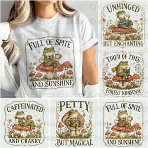 May include: A white t-shirt featuring a graphic of a frog sitting on a mushroom, holding a cup, with the text "FULL OF SPITE AND SUNSHINE" enclosed in a decorative border. The design includes surrounding wildflowers and smaller mushrooms.