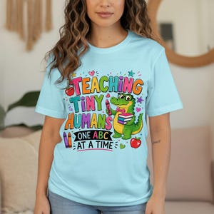 May include: A light blue t-shirt featuring a colorful graphic of a green alligator wearing glasses and a bow tie, holding a stack of books. The text reads "TEACHING TINY HUMANS ONE ABC AT A TIME" with illustrations of crayons, apples, and hearts.