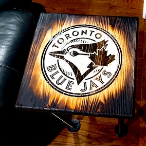 Op de afbeelding: Vierkante houten bijzettafel met het logo van de Toronto Blue Jays erin gebrand. Het logo, een witte cirkel met een blauwe gaai en tekst, is in het hout gebrand. De tafel heeft een donkere, gebrande afwerking en zwarte metalen poten. Ongeveer 40 cm x 40 cm.