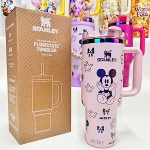 May include: A light pink Stanley Quencher H2.0 FlowState tumbler with a 40 oz capacity, featuring a Mickey Mouse graphic, bows, and hand prints. The tumbler includes a handle, a clear lid with a straw, and comes in a brown cardboard box.