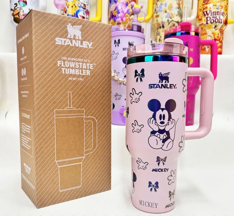 May include: A light pink Stanley Quencher H2.0 FlowState tumbler with a 40 oz capacity, featuring a Mickey Mouse graphic, bows, and hand prints. The tumbler includes a handle, a clear lid with a straw, and comes in a brown cardboard box.