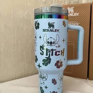 May include: A light blue Stanley tumbler featuring a rainbow metallic rim and handle. The body is decorated with a repeating pattern of Stitch characters, hibiscus flowers, and star shapes. The word Stitch is printed in a rainbow metallic font.