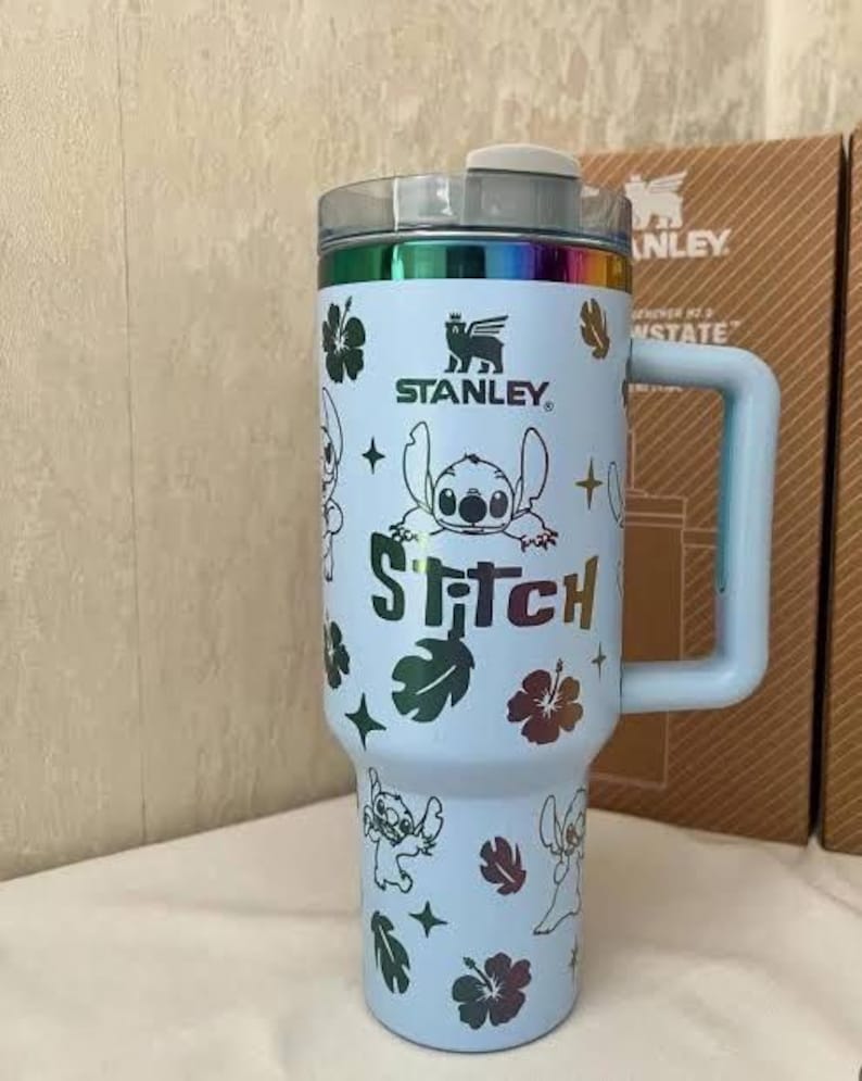 May include: A light blue Stanley tumbler featuring a rainbow metallic rim and handle. The body is decorated with a repeating pattern of Stitch characters, hibiscus flowers, and star shapes. The word Stitch is printed in a rainbow metallic font.