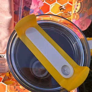 May include: A circular tumbler lid featuring a bright yellow plastic handle with a white center insert and a small circular opening. The lid is positioned over a container decorated with a colorful honeycomb pattern and cartoon characters.