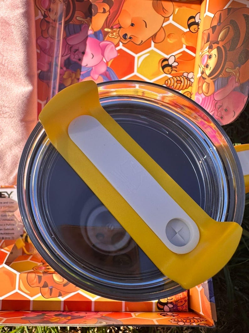 May include: A circular tumbler lid featuring a bright yellow plastic handle with a white center insert and a small circular opening. The lid is positioned over a container decorated with a colorful honeycomb pattern and cartoon characters.