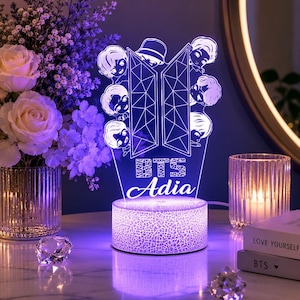 Może przedstawiać: Lampa LED z przezroczystego akrylu z geometrycznym logo BTS, ilustracjami postaci z kreskówek oraz wygrawerowanym napisem "BTS Adia". Lampa stoi na okrągłej, teksturowanej podstawie, która emituje fioletowe światło.