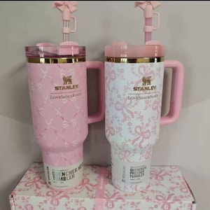 May include: Two Stanley Quencher tumblers with handles and straw toppers. The left tumbler is pink with a white ribbon pattern, and the right is white with a pink ribbon pattern. Both feature gold Stanley LoveShackFancy branding and lids.