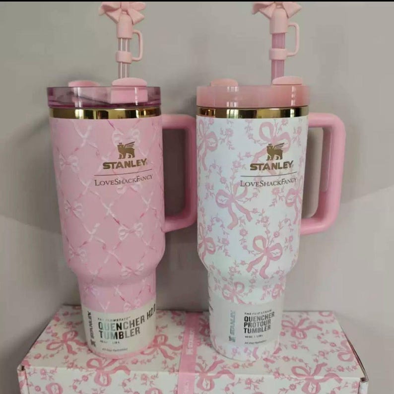 May include: Two Stanley Quencher tumblers with handles and straw toppers. The left tumbler is pink with a white ribbon pattern, and the right is white with a pink ribbon pattern. Both feature gold Stanley LoveShackFancy branding and lids.