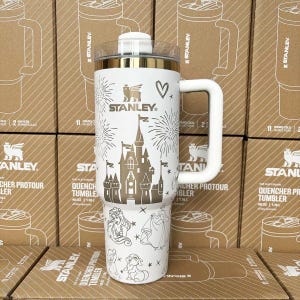 May include: A white 40-ounce Stanley Quencher tumbler featuring a gold-colored castle graphic, fireworks, and various character illustrations. The tumbler has a clear lid, a white handle, and the Stanley logo printed in gold on the front surface.
