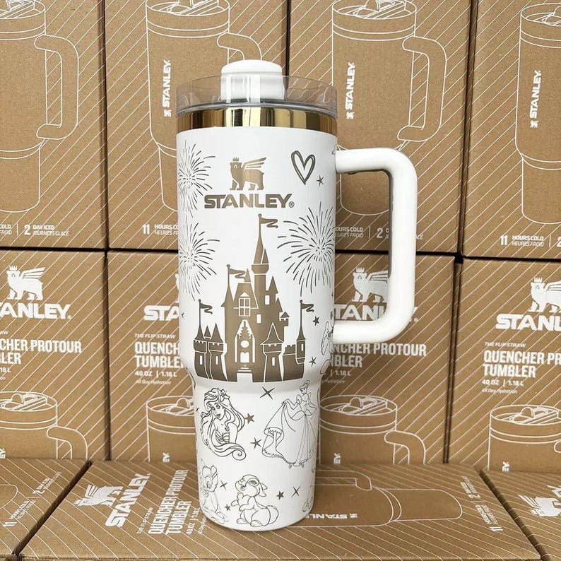 May include: A white 40-ounce Stanley Quencher tumbler featuring a gold-colored castle graphic, fireworks, and various character illustrations. The tumbler has a clear lid, a white handle, and the Stanley logo printed in gold on the front surface.