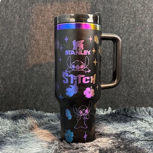 May include: A matte black Stanley tumbler with a handle and a lid featuring an iridescent rainbow finish. The cup is decorated with holographic graphics of the character Stitch, hibiscus flowers, and stars, with the text Stanley and Stitch.