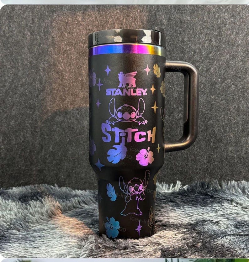 May include: A matte black Stanley tumbler with a handle and a lid featuring an iridescent rainbow finish. The cup is decorated with holographic graphics of the character Stitch, hibiscus flowers, and stars, with the text Stanley and Stitch.