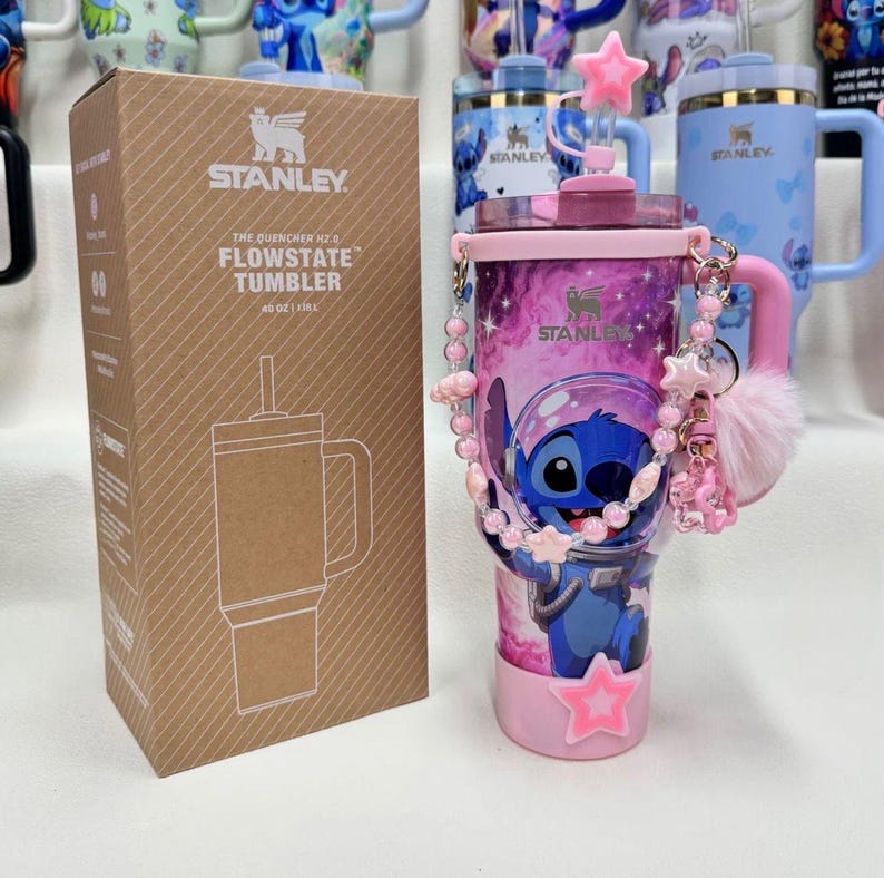 May include: A 40 oz Stanley FlowState tumbler in a pink and purple galaxy print featuring a Stitch character graphic. The tumbler includes a pink handle, a straw with a star topper, a beaded charm strap, a pink pom-pom, and its original box.