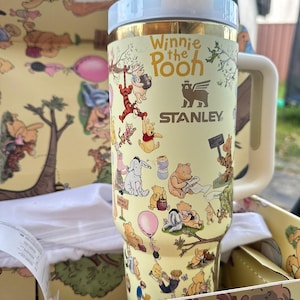 May include: A yellow Stanley travel mug featuring Winnie the Pooh characters. The mug has a light blue lid and handle, with the words "Winnie the Pooh" printed on the side. The mug is in a matching yellow box with Winnie the Pooh illustrations.
