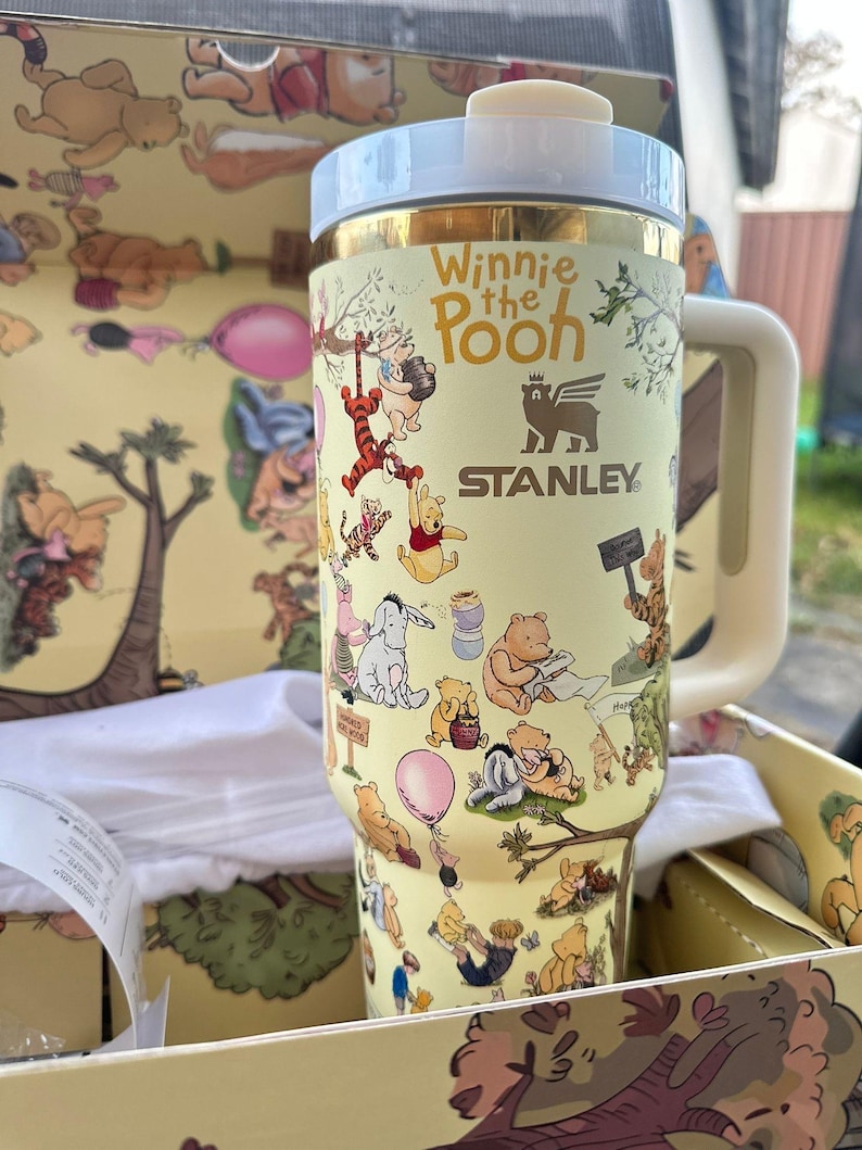 May include: A yellow Stanley travel mug featuring Winnie the Pooh characters. The mug has a light blue lid and handle, with the words "Winnie the Pooh" printed on the side. The mug is in a matching yellow box with Winnie the Pooh illustrations.