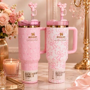 May include: Two 40 oz Stanley LoveShackFancy Quencher H2.0 tumblers with pink and white bow patterns. Each features a gold-tone rim, a pink handle, a clear lid, and a pink straw topped with a small bow accessory. The base labels read 40 oz | 1.18 L.