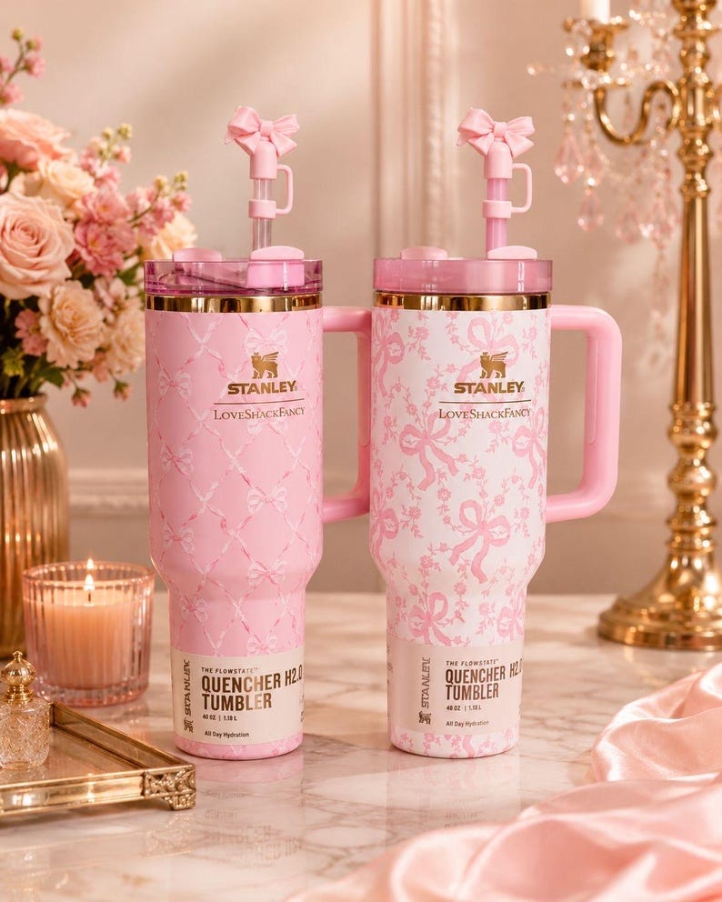 May include: Two 40 oz Stanley LoveShackFancy Quencher H2.0 tumblers with pink and white bow patterns. Each features a gold-tone rim, a pink handle, a clear lid, and a pink straw topped with a small bow accessory. The base labels read 40 oz | 1.18 L.