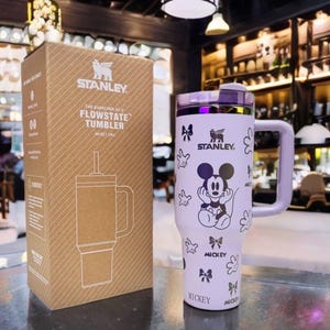 May include: A light purple Stanley Flowstate tumbler with a Mickey Mouse graphic, featuring a handle, a lid, and a straw. The tumbler is displayed next to its brown cardboard packaging, which is printed with the Stanley logo and product name.